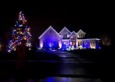 Large brick home at night decorated with cool white roofline lights and blue accent lighting on shrubs, with a tall outdoor Christmas tree glowing in multicolored lights on the left side of the yard.